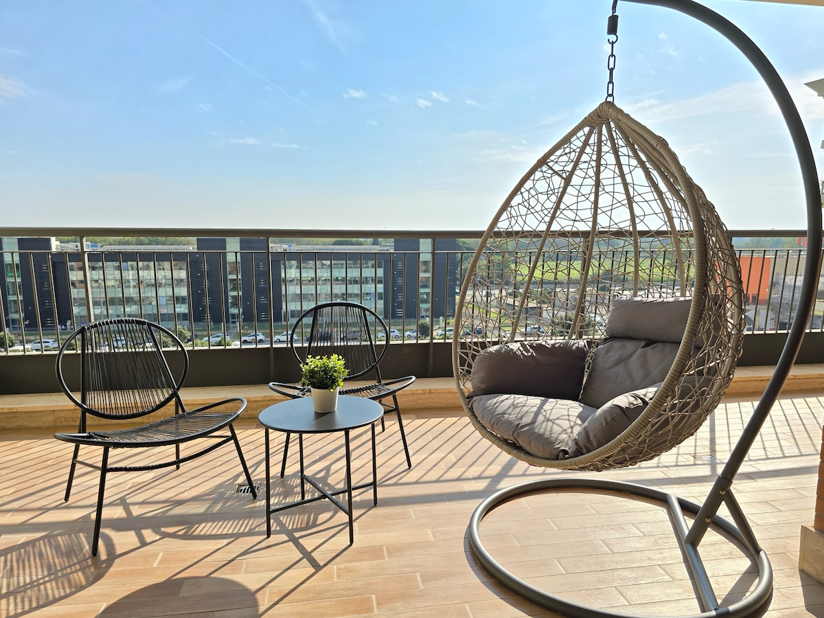 A modern terrace features a hanging chair accompanied by two black wire chairs and a small table. A potted plant sits atop the table. The railing offers unobstructed views of the surroundings, bathed in natural light.