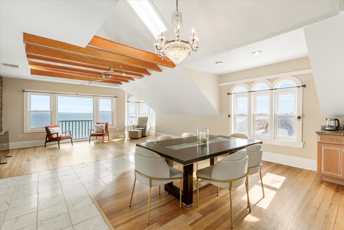 A spacious dining area is illuminated by natural light streaming through three large windows. A modern table with seating for six is centered beneath an elegant chandelier, complemented by comfortable armchairs positioned nearby. Expansive ocean views are visible through the windows.