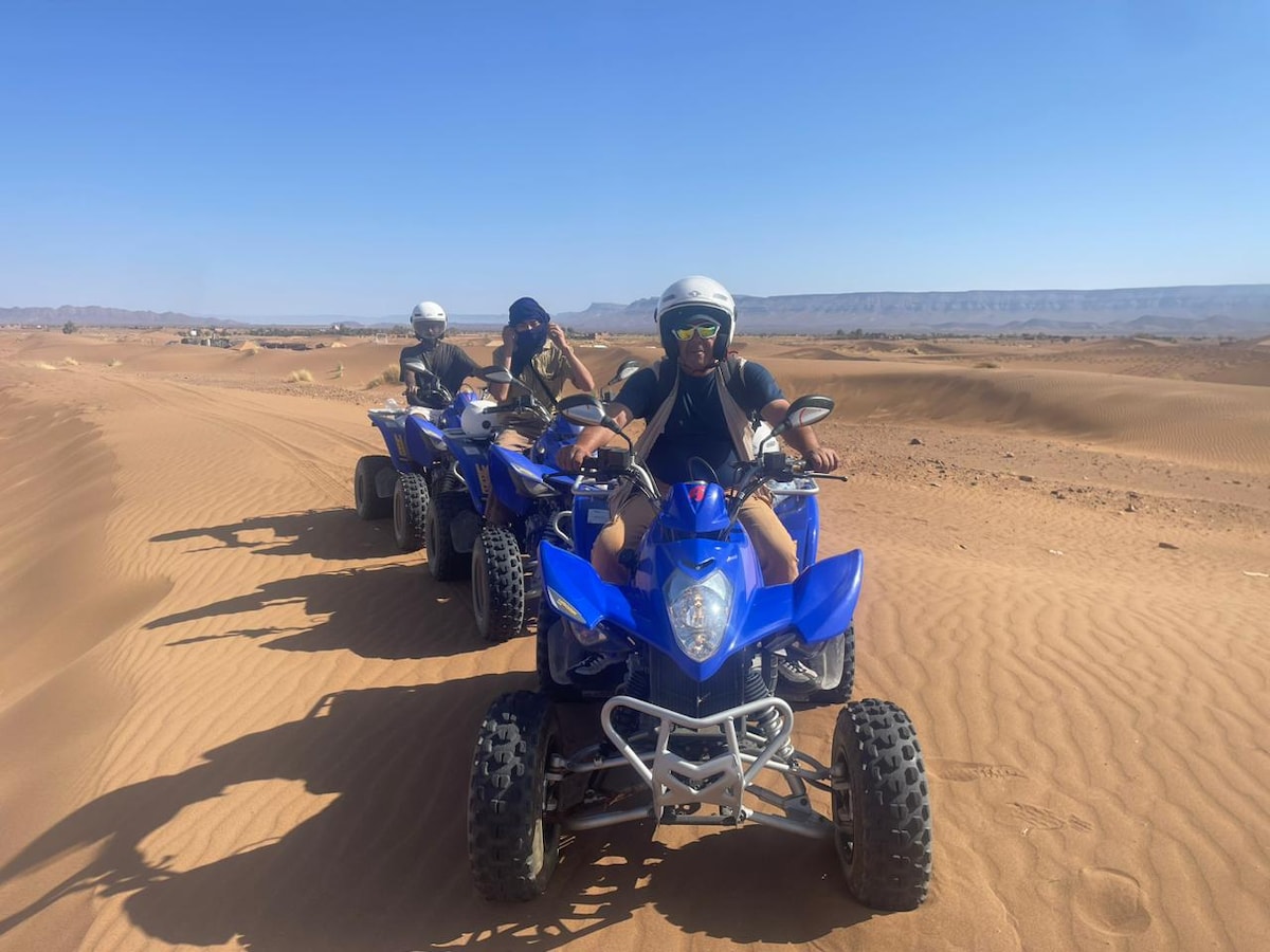 Three individuals are positioned on blue quad bikes in a desert landscape, with rolling sand dunes extending into the distance. The sun casts bright light over the scene, highlighting the textures of the sandy terrain and the performance vehicles.