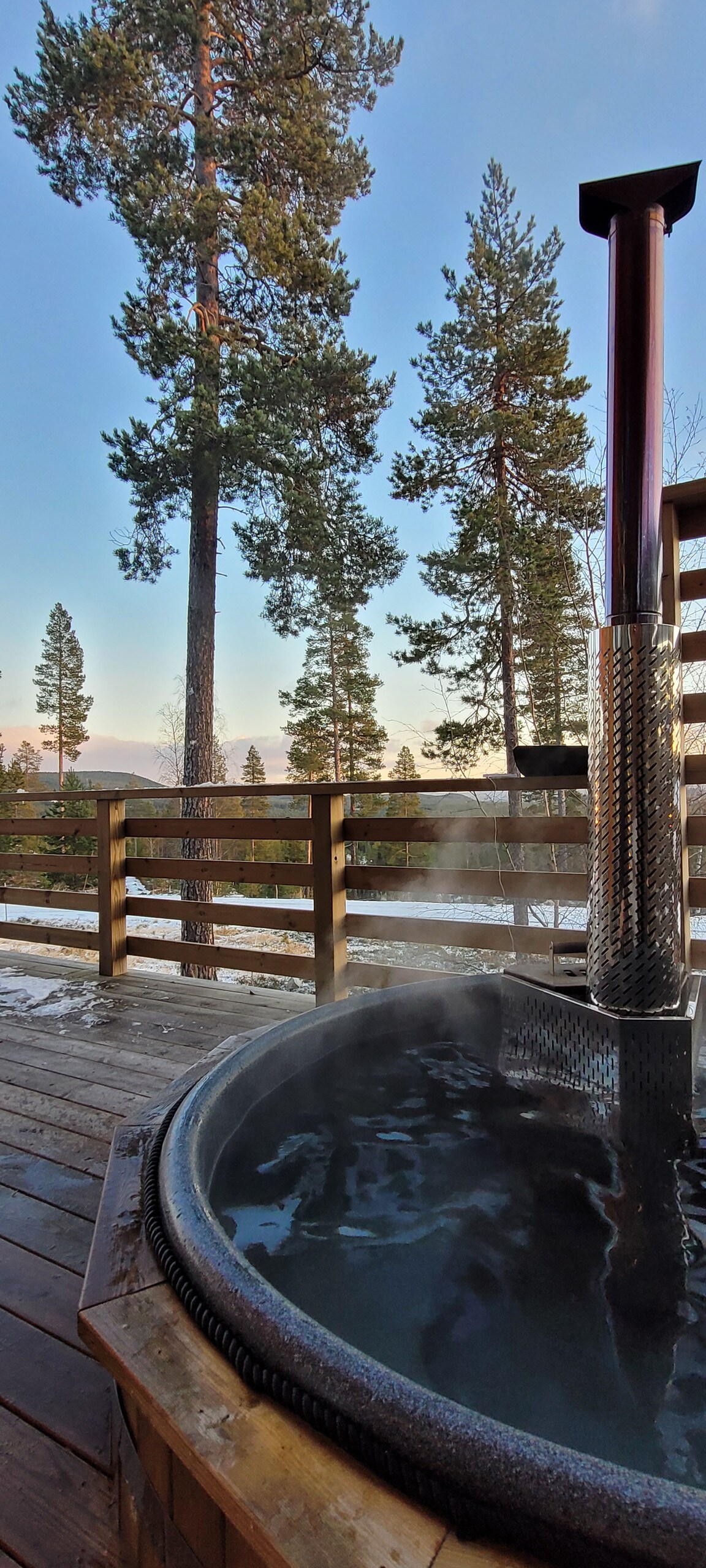A hot tub with steam rising sits on a wooden deck, surrounded by tall trees. The view beyond includes mountains and a clear sky, indicating a tranquil outdoor setting. The wooden railing enhances the rustic charm of the area.