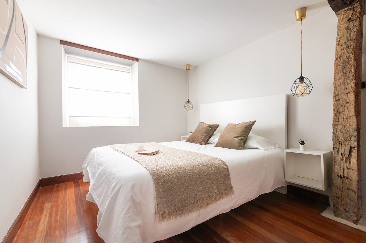 A bright sleeping area features a spacious bed with light linens and two decorative pillows. The room is complemented by warm wooden flooring and a window allowing natural light to enter. Pendant lamps hang from the ceiling, enhancing the modern design.
