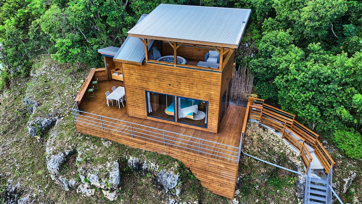 The lodge is nestled among lush greenery, constructed with wooden materials to blend with nature. Large glass doors create a seamless transition between indoor and outdoor spaces. The deck features seating areas and a dining table, offering expansive views of the surrounding landscape.