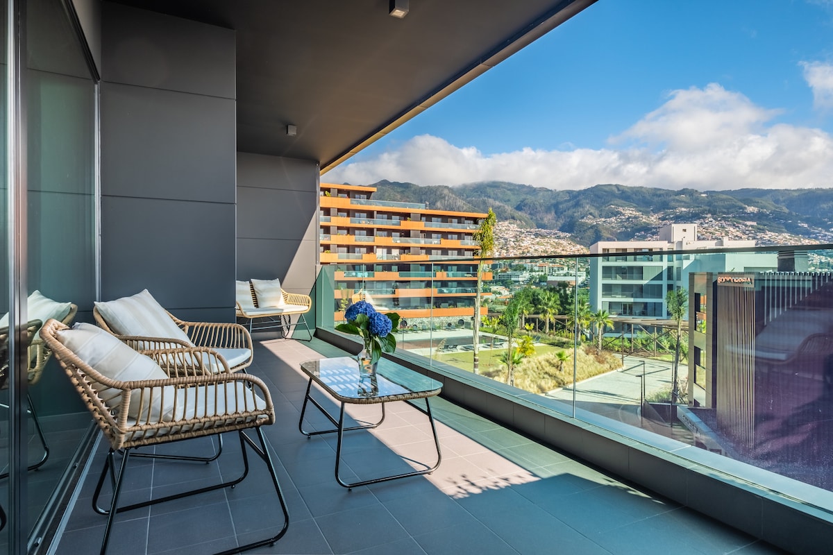 A modern balcony offers seating with wicker chairs and a glass table, surrounded by views of the city and mountains. The area is brightened by natural light, creating an inviting outdoor space. Lush greenery is visible in the distance, enhancing the serene atmosphere.