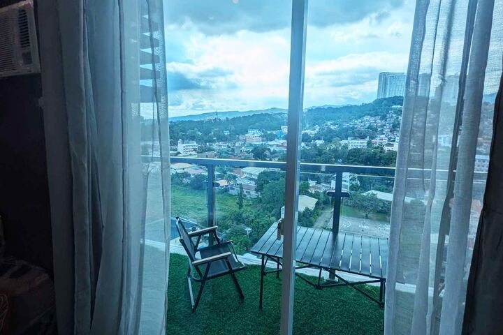 A view from the balcony showcases a city landscape framed by translucent curtains. A small table and two chairs are positioned on a grassy floor, offering an inviting outdoor space to enjoy the surroundings. The mountains are visible in the distance under a cloudy sky.