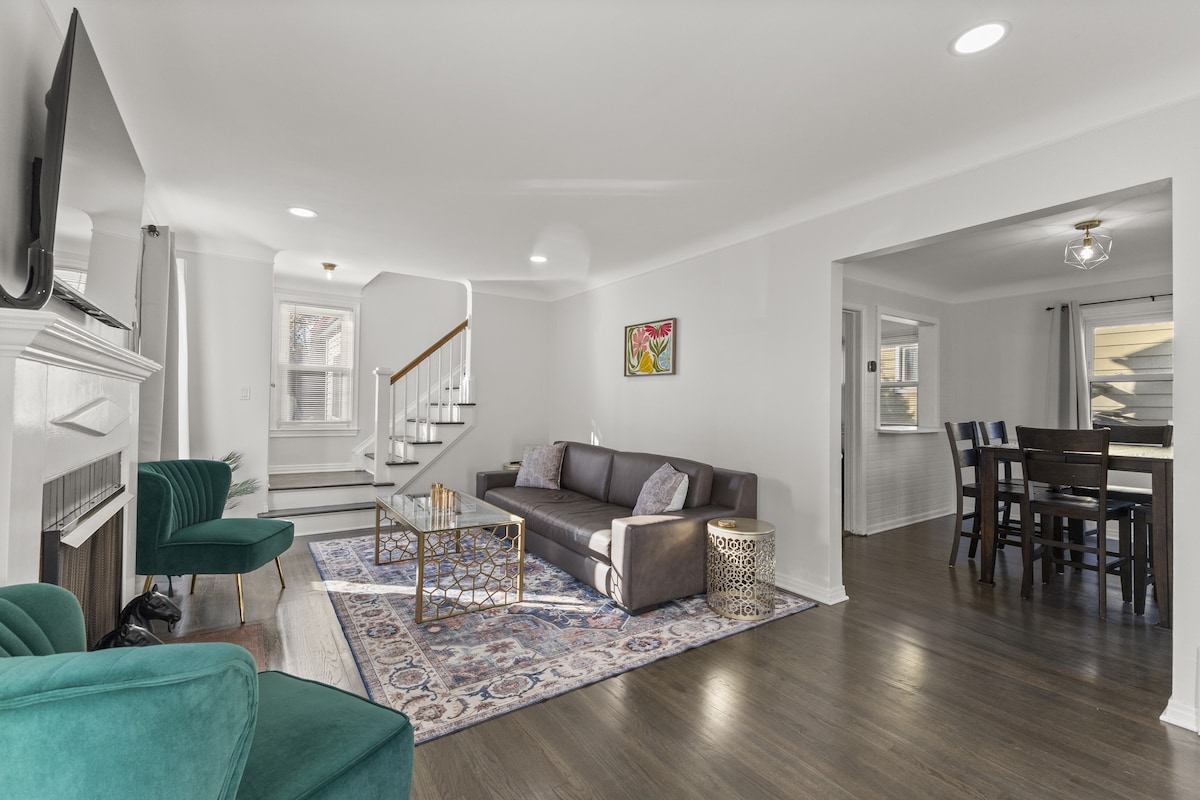 A modern living area features a dark gray sofa paired with two green chairs. A gold coffee table is placed on a decorative area rug. A television is mounted above a white fireplace, and a staircase is visible in the background, leading to an upper level.