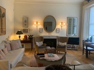 The spacious drawing room features a light-colored sofa, two comfortable armchairs, and a modern coffee table. Soft lighting is provided by wall sconces and a lamp, creating a cozy atmosphere. A large mirror and artwork adorn the walls, and a small dining area is visible in the background.