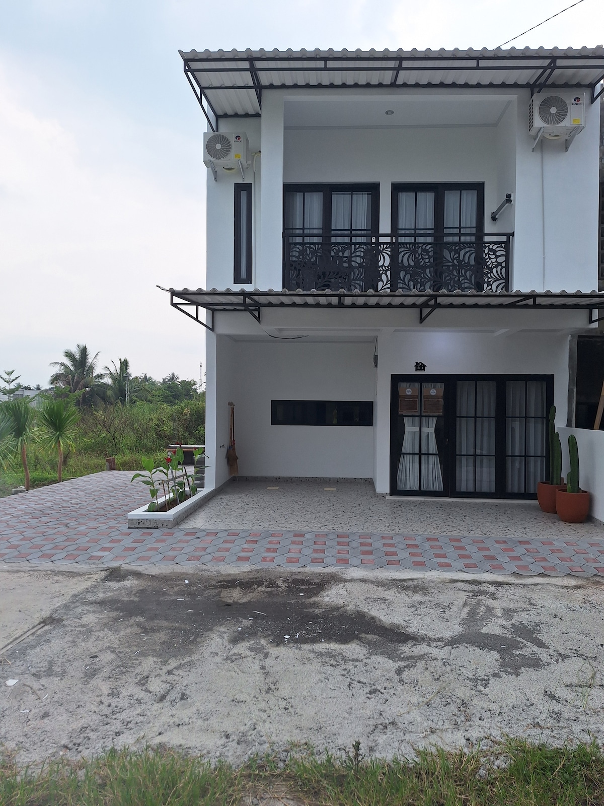 A two-story building features a modern façade with large black-framed windows and a decorative balcony. The ground level includes a covered entrance with a pathway made of patterned pavers. Surrounding greenery and palm trees add a serene backdrop.