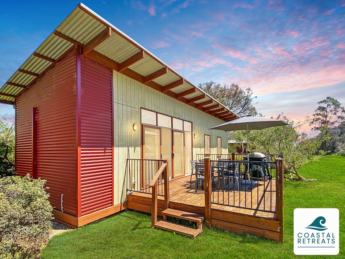 The exterior of Moya's Cottage is depicted, showcasing a modern design with a combination of wooden and metal surfaces. A spacious deck is visible, featuring an outdoor table, chairs, and an umbrella for shade. Lush green grass surrounds the cottage, enhancing its inviting appearance.