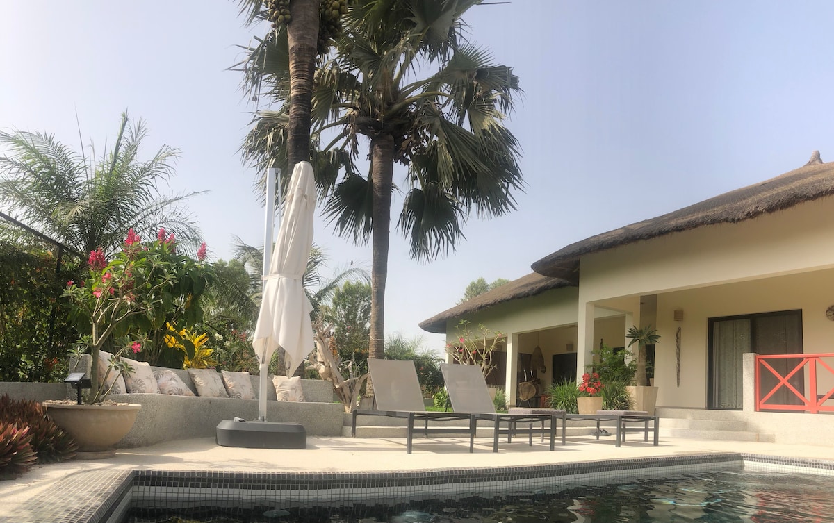 A spacious outdoor area features sun loungers arranged near a tranquil pool. Lush tropical plants surround the space, complemented by vibrant flowers. A large umbrella provides shade, while a charming thatched-roof structure serves as a backdrop, creating a relaxing atmosphere for guests.