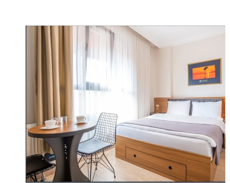 A well-lit room features a spacious bed with crisp white linens. A small round table with two chairs is positioned nearby, accompanied by breakfast items. Sheer curtains allow soft light to filter in, enhancing the serene atmosphere.