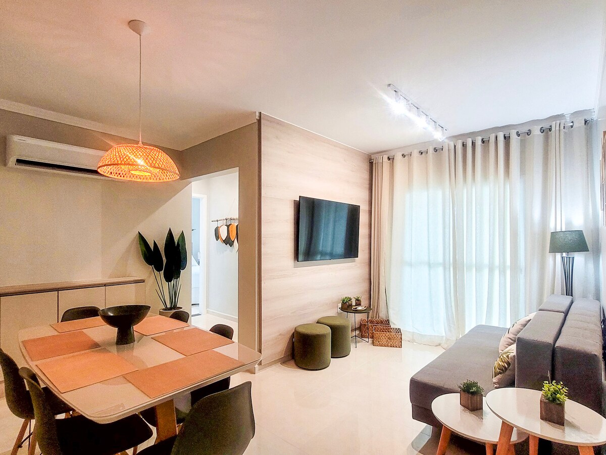 A well-lit living area features a comfortable gray sofa and a dining table set for six. A large television is mounted on the wall, while light drapes softly cover the windows. Decorative plants add a touch of greenery to the space.