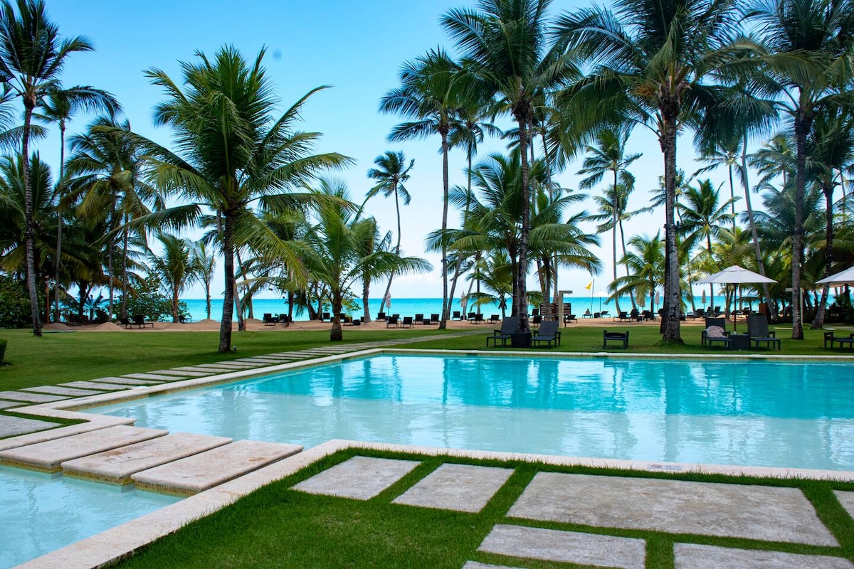 A serene pool area is framed by lush greenery and swaying palm trees, with lounge chairs positioned along the edges. The clear blue water reflects the sky, while the beach is visible in the background, creating a tranquil outdoor atmosphere.