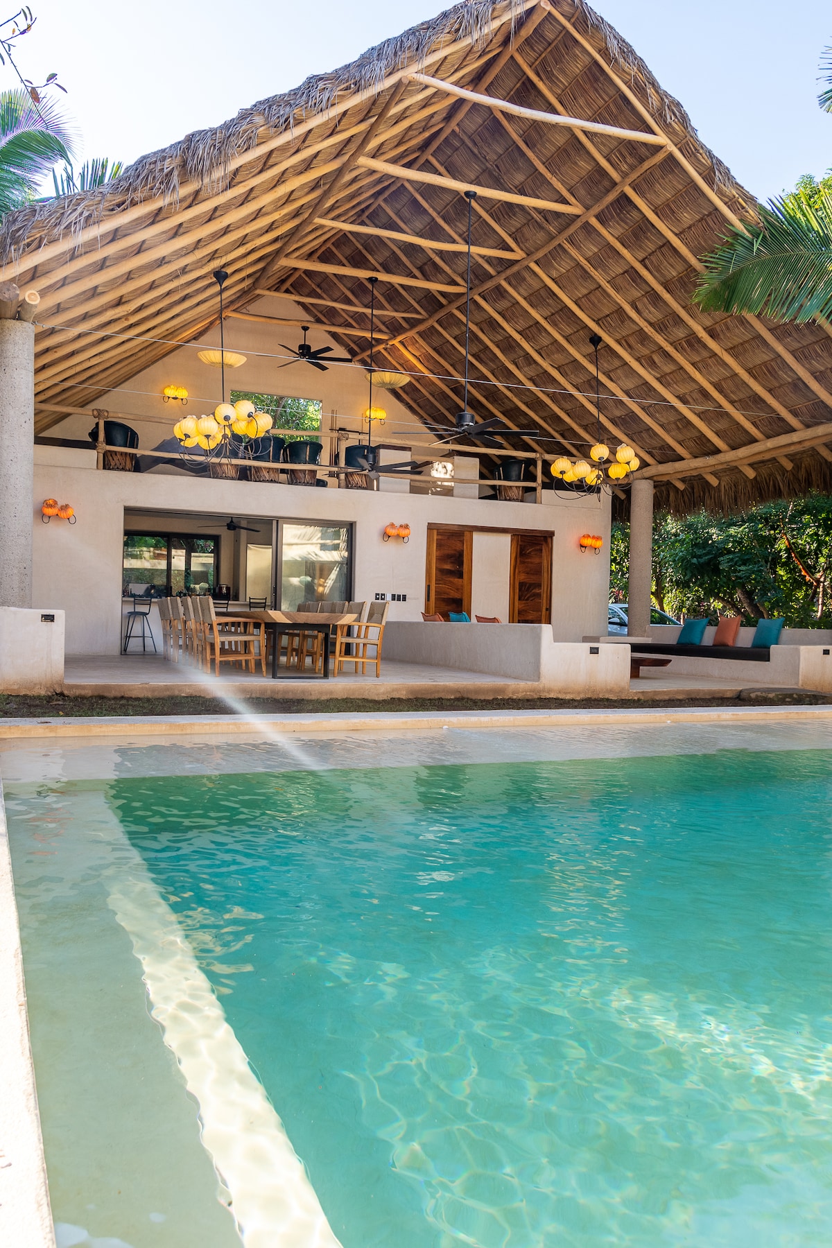 An open-concept space is highlighted by a high thatched roof. A large swimming pool reflects the natural surroundings, while a dining area with wooden furnishings is visible in the background. Soft lighting fixtures hang gracefully, enhancing the villa's light and airy feel.