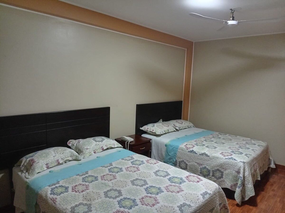 A welcoming room features two comfortable individual beds, each topped with patterned covers. A bedside table is positioned between the beds, and wall-mounted fixtures are present. Neutral-toned walls and wooden flooring contribute to a cozy atmosphere, complemented by a ceiling fan for added ventilation.