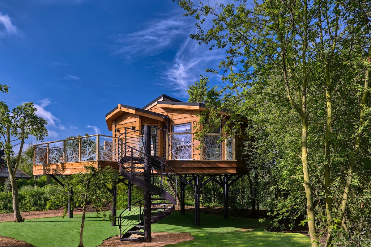 A modern treehouse with a wooden exterior is elevated above the ground, supported by a spiral staircase. Large windows allow natural light to fill the interior. Surrounding greenery creates a natural setting, while a clear blue sky adds to the serene atmosphere.
