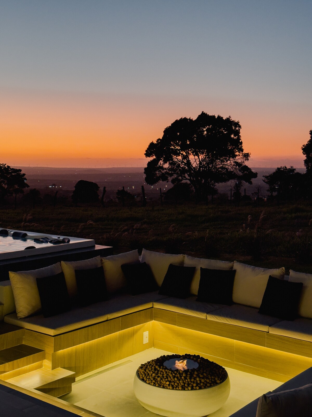 An outdoor seating area features a spacious L-shaped couch adorned with dark pillows. A circular fire pit is set in the center, surrounded by smooth decorative stones, as a vibrant sunset illuminates the sky in warm hues. Expansive views of the landscape are visible beyond.