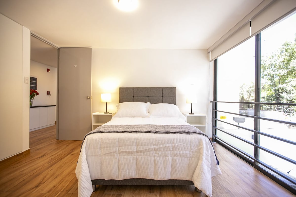 A bright sleeping area features a spacious bed with crisp white linens and soft gray accents. Two bedside lamps provide gentle illumination, and large windows invite natural light while offering views of the surrounding greenery.