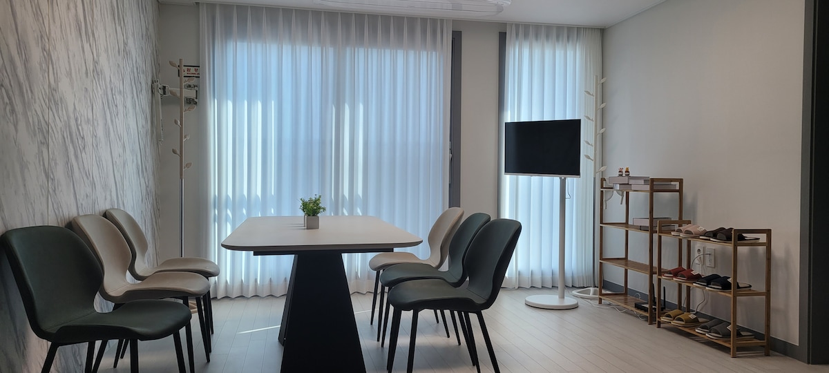 A bright meeting space features large windows covered with sheer curtains, allowing natural light to filter through. A central white table is surrounded by six comfortable chairs. A television is mounted on a stand, and a small plant decorates the table, adding a touch of greenery.