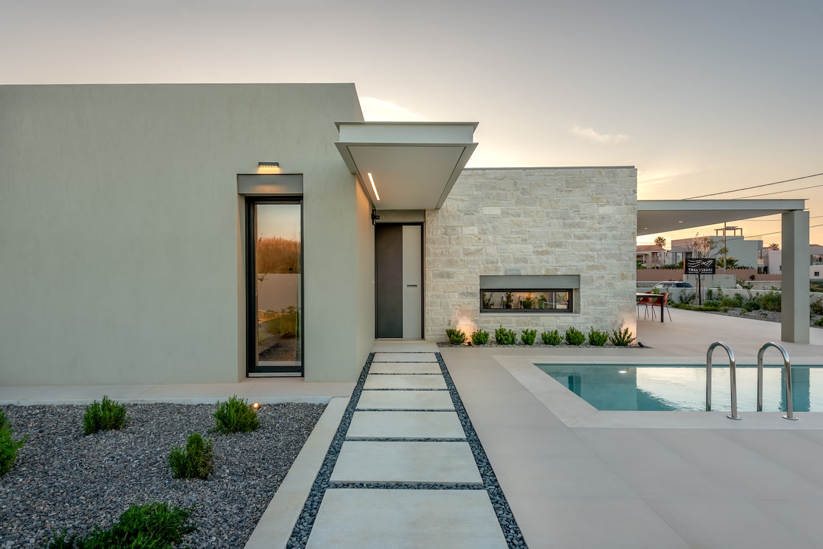 The entrance to the villa is framed by modern stone and stucco walls. A smooth pathway leads to the front door, accompanied by landscaped gardens featuring neatly arranged rocks and low greenery. The private swimming pool is visible to the right, providing a serene backdrop.