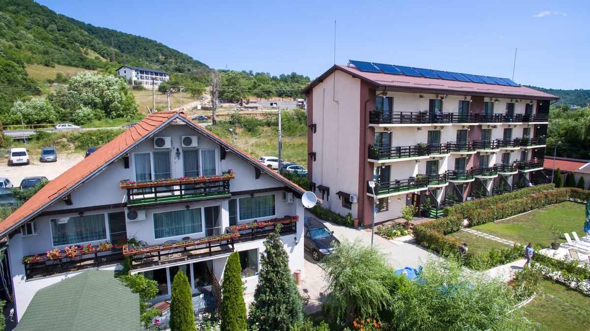 The exterior of Danubia Maison showcases a two-building layout. The left structure features a welcoming design with balconies adorned with greenery, while the right structure has multiple levels and solar panels on the roof. A landscaped area with ample parking surrounds the buildings.