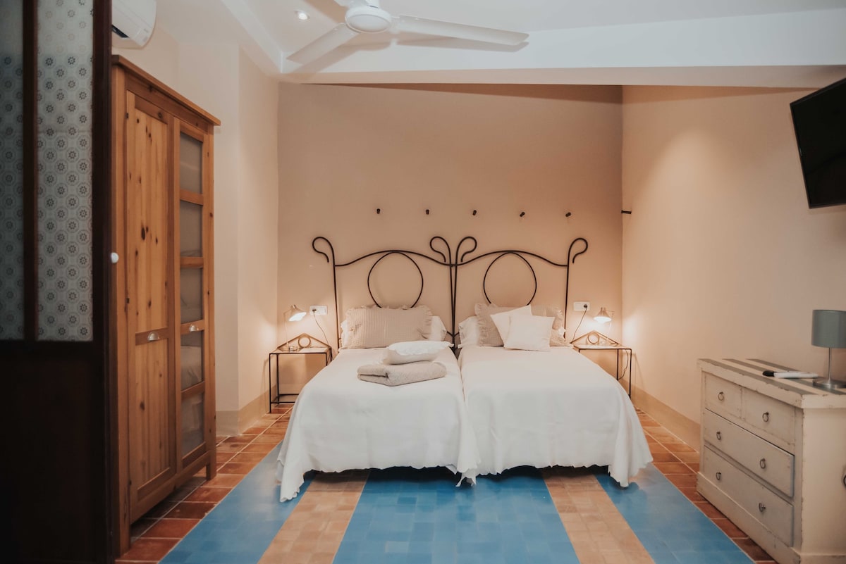 A welcoming room features two spacious twin beds with elegant wrought iron headboards. Soft white linens are neatly arranged, and light from bedside lamps casts a warm glow. A wooden wardrobe and a small dresser provide storage, enhancing the room's functionality.
