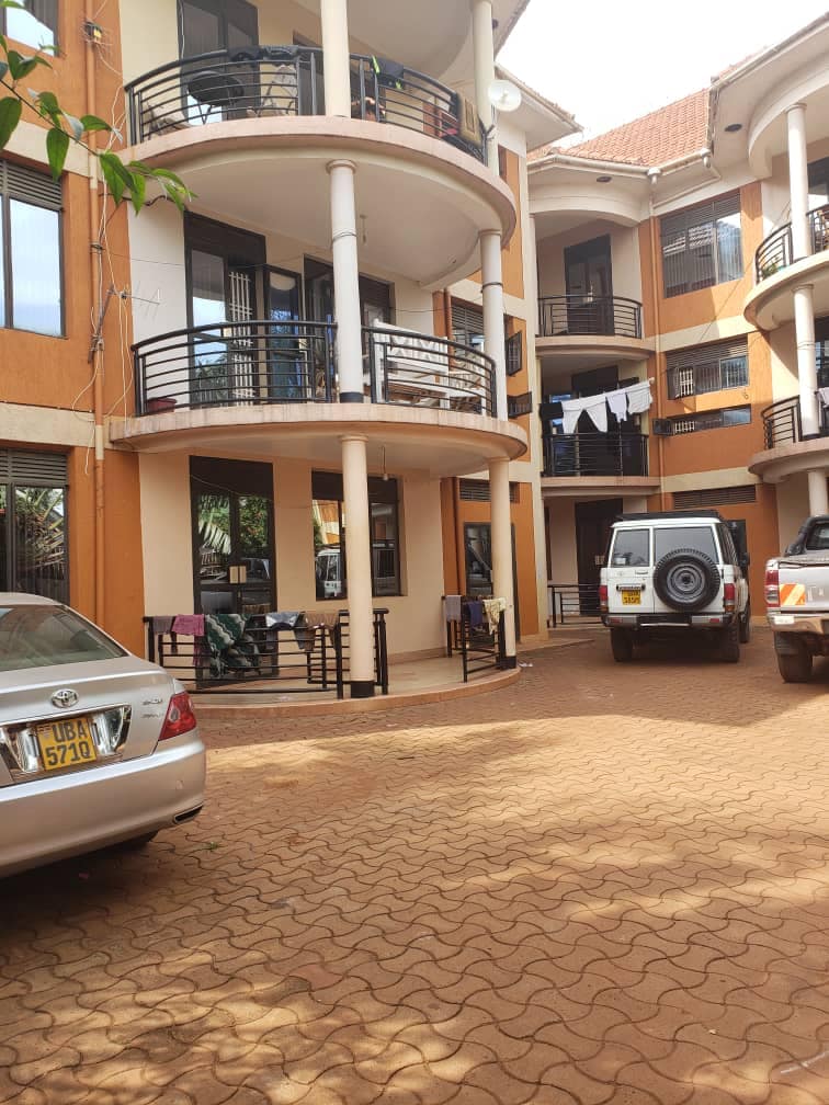 A courtyard is shown with multiple vehicles parked, surrounded by a multi-story building featuring balconies. Clothes are hanging on a clothesline, and a variety of greenery is visible near the ground level. The paved ground is marked by a patterned layout.