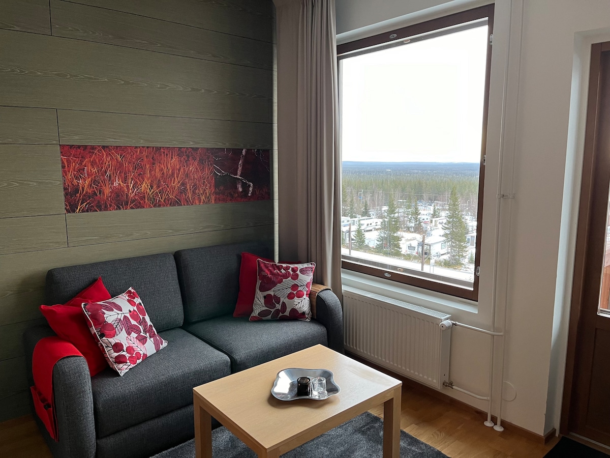 The living area features a comfortable gray sofa adorned with red and white floral cushions. A light wooden coffee table is positioned in front. A large window offers a view of the surrounding landscape, with distant trees and open spaces visible.