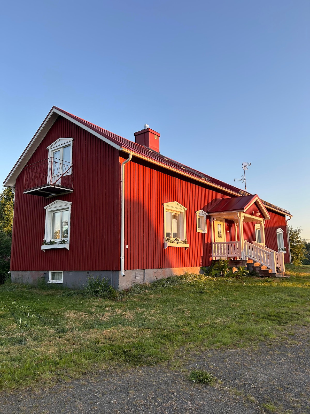 The charming red house features a classic exterior with white trim, highlighting multiple windows and a small balcony. A welcoming front porch leads to the entrance, surrounded by green grass and a clear blue sky, emphasizing the serene countryside setting.