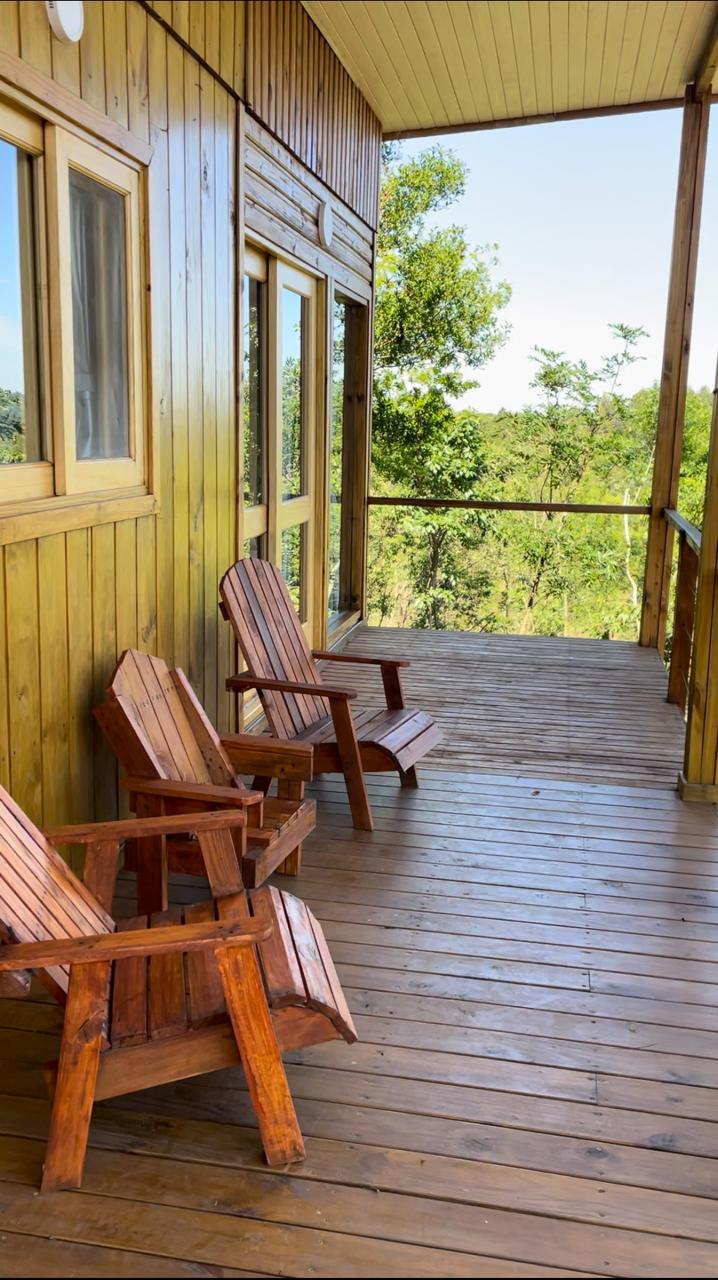A wooden balcony features two rustic chairs crafted from dark wood. The floor is made of polished planks, and large windows provide views of the surrounding greenery. Sunlight brightens the space, creating a serene atmosphere.