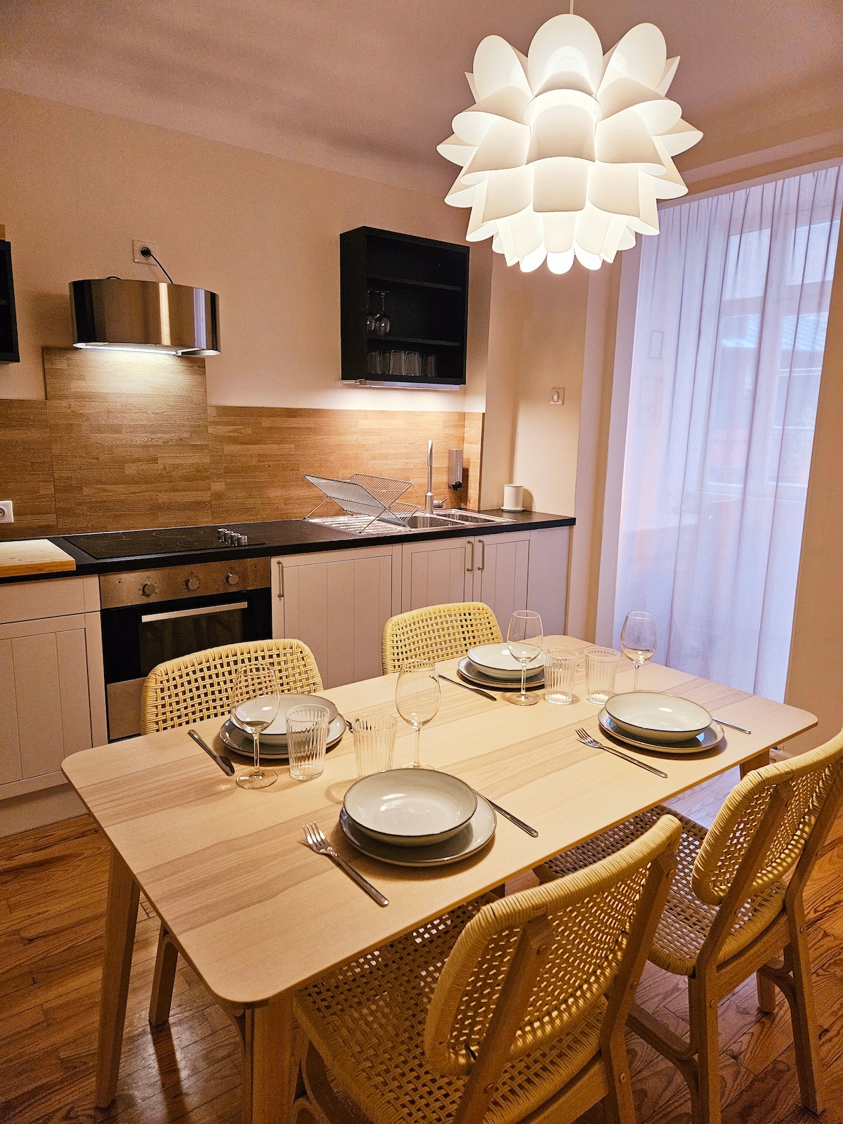 A dining area is presented with a light wooden table set for four, featuring plates, glasses, and silverware. A stylish pendant light hangs above the table, while black countertops and wood paneling enhance the kitchen's atmosphere. Natural light filters through a sheer curtain.