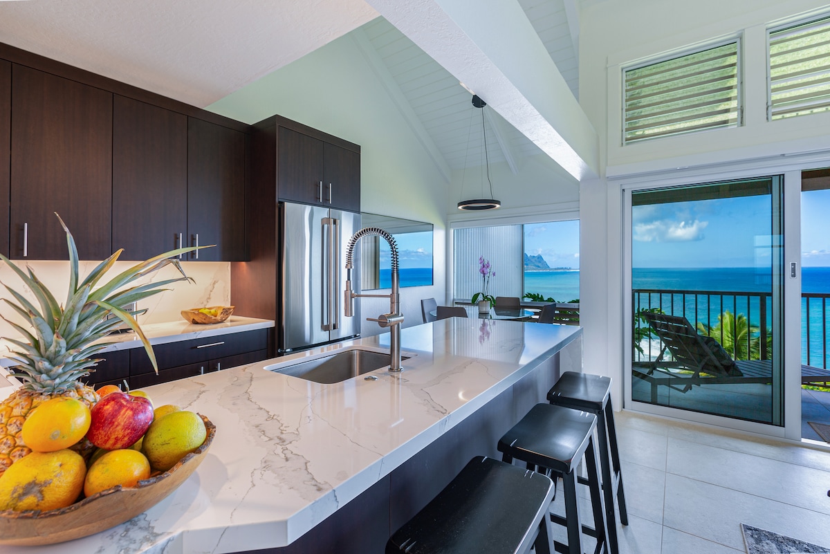 A modern kitchen is highlighted by sleek dark cabinetry and a marble countertop featuring a bowl of fresh fruit. It opens to a panoramic view of the ocean through large sliding glass doors, with natural light illuminating the space.