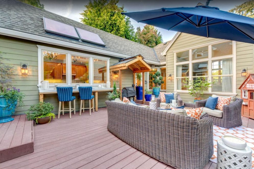 An enclosed patio area features comfortable wicker seating, complemented by decorative cushions and a large umbrella for shade. A rustic bar counter with high stools invites casual dining. Lush plants and a children's playhouse enhance the inviting outdoor space, perfect for gatherings.