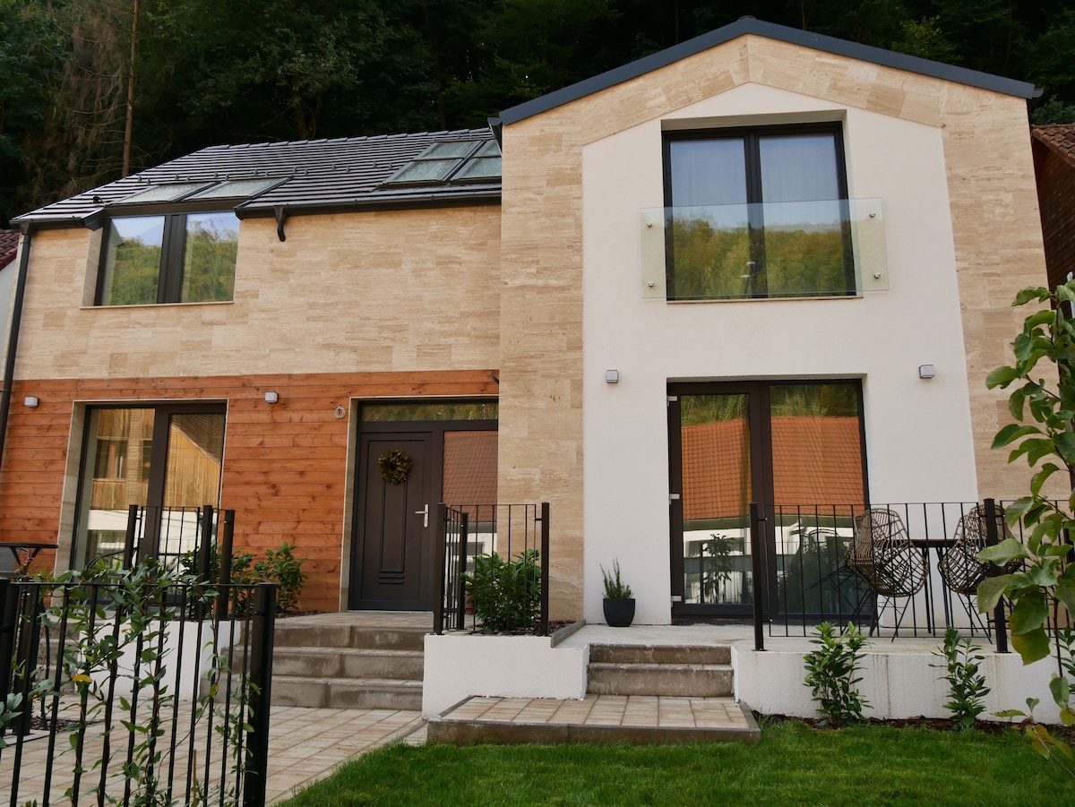 The exterior of the modern two-story building features a blend of natural stone and wooden accents. Large windows allow ample light, and a welcoming entryway is complemented by decorative plants. A manicured lawn and stone pathway lead to the main entrance.