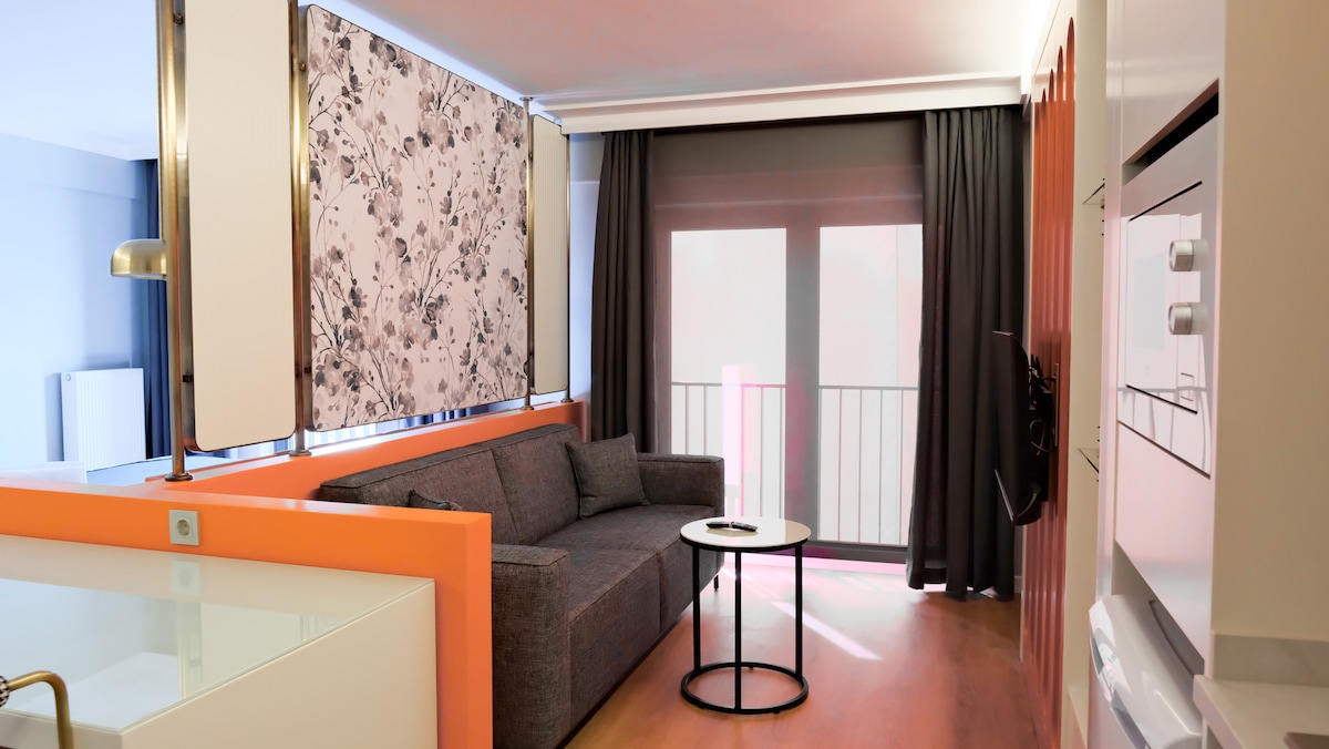 A modern living space features a gray sofa paired with a black round coffee table. Large windows are adorned with floral-patterned curtains and solid dark drapes, allowing light to filter through. The open layout is enhanced by a combination of orange and neutral-colored walls.
