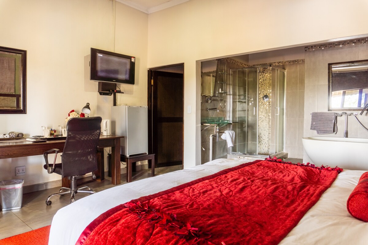 A spacious bedroom features a large bed covered with red linens, complemented by a modern desk and chair. A flat-screen television is mounted on the wall, while a glass-enclosed shower is visible beside a standalone bathtub. A refrigerator is placed near the entrance.