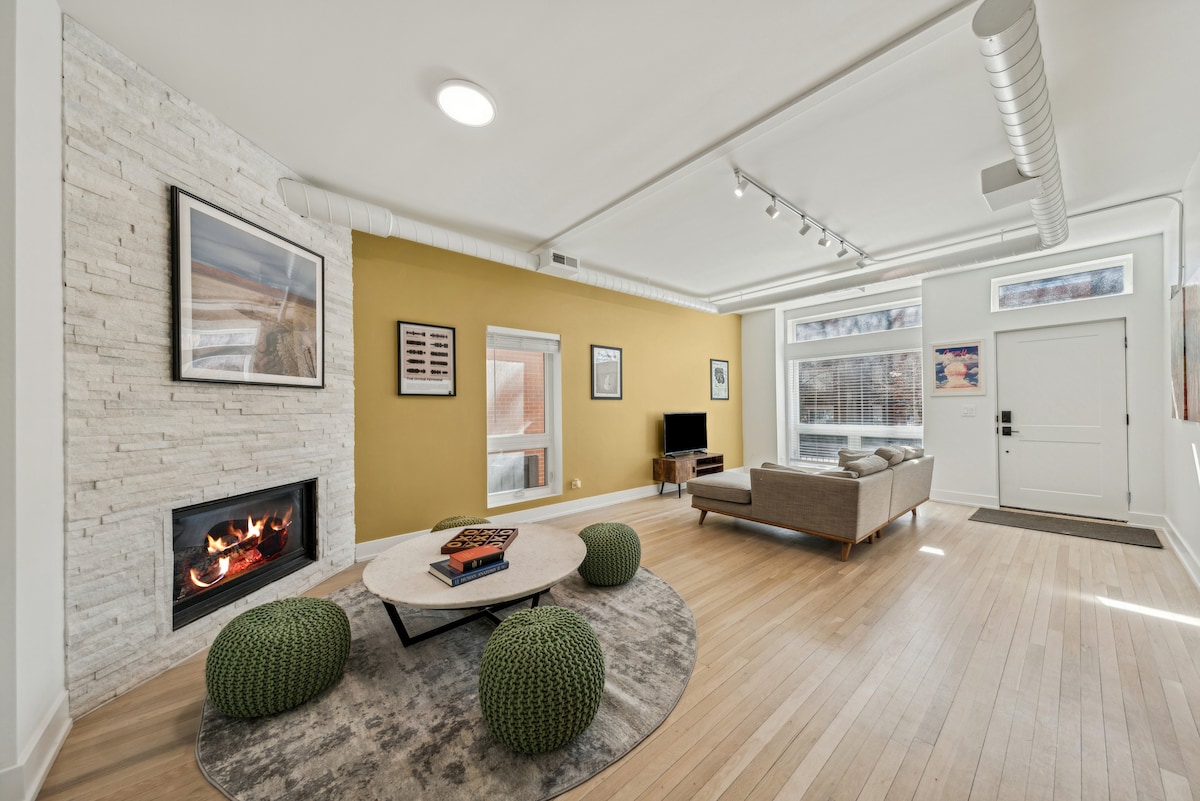 An open-concept living area features light wood flooring and a stone accent wall. A modern sofa is positioned near a coffee table on a patterned rug. A flat-screen television is mounted on the wall, and natural light fills the space through large windows.