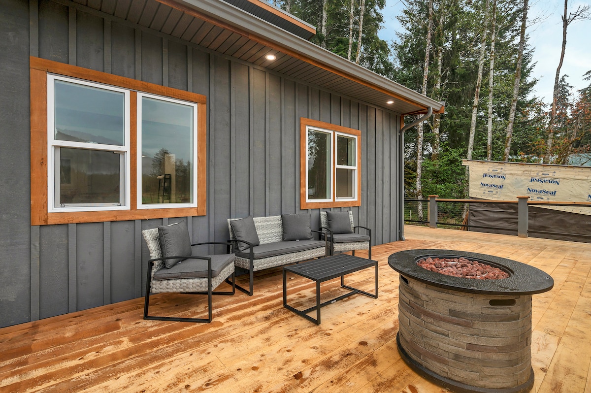 A furnished deck area features a circular stone fire pit surrounded by a wooden floor. Two comfortable chairs and a matching sofa are positioned near large windows, providing views of the surrounding trees.