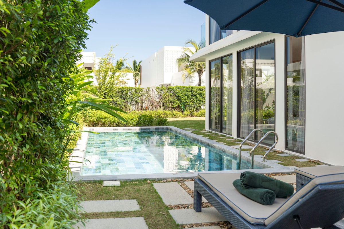 A private pool is surrounded by lush greenery, showcasing clear water reflecting sunlight. Comfortable lounge chairs are placed nearby, with towels neatly rolled at the foot. Large glass doors offer a view into the villa, enhancing the connection between indoor and outdoor spaces.