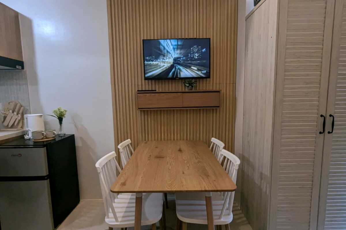 A dining area is presented with a wooden table surrounded by four white chairs. A wall-mounted television is positioned above a wooden shelf, contrasting with the textured wood paneling behind. A small flower arrangement adds a touch of greenery to the space.