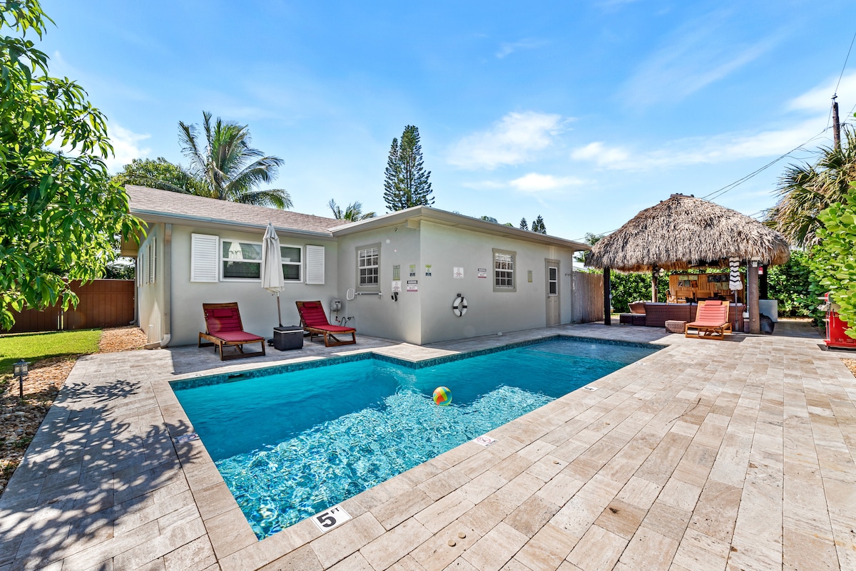A heated saltwater pool is featured, surrounded by a spacious patio crafted with stone tiles. Lounge chairs in vibrant red are positioned nearby, complemented by a thatched tiki hut equipped with seating and a bar. Lush greenery enhances the tropical ambiance of the backyard.