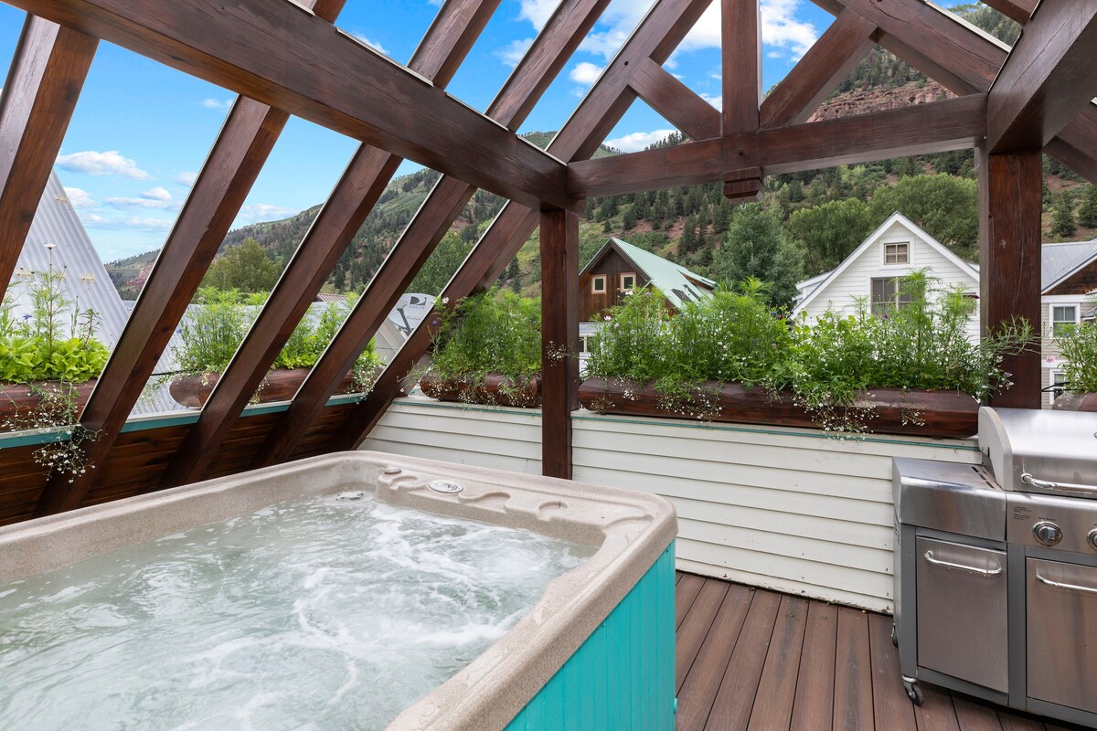 An outdoor hot tub is positioned on a wooden deck beneath a vaulted wooden structure, surrounded by lush greenery in planter boxes. A grill is visible nearby, with scenic mountain views framed by the clear sky above.