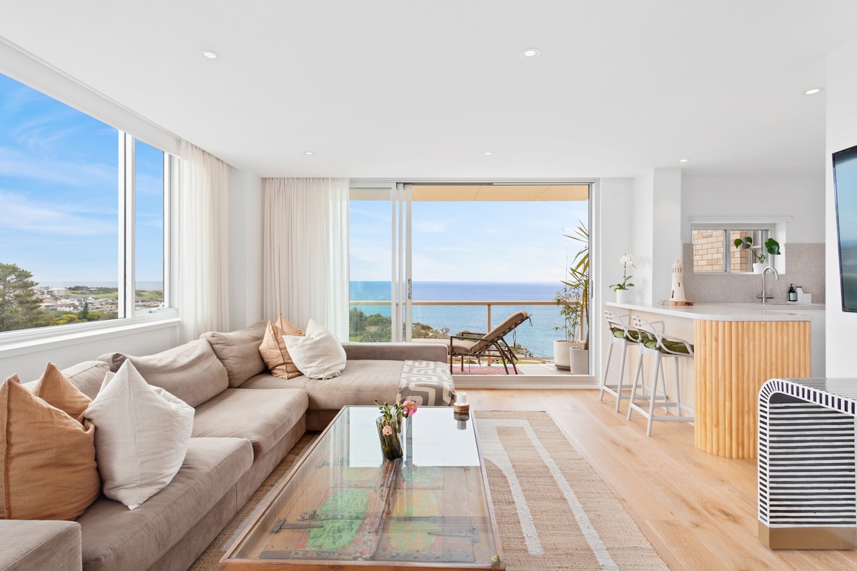 A bright living area features a spacious sectional sofa adorned with decorative pillows, facing large windows that offer expansive ocean views. A glass coffee table is centered on a light area rug, while a modern kitchen bar area is visible in the background.