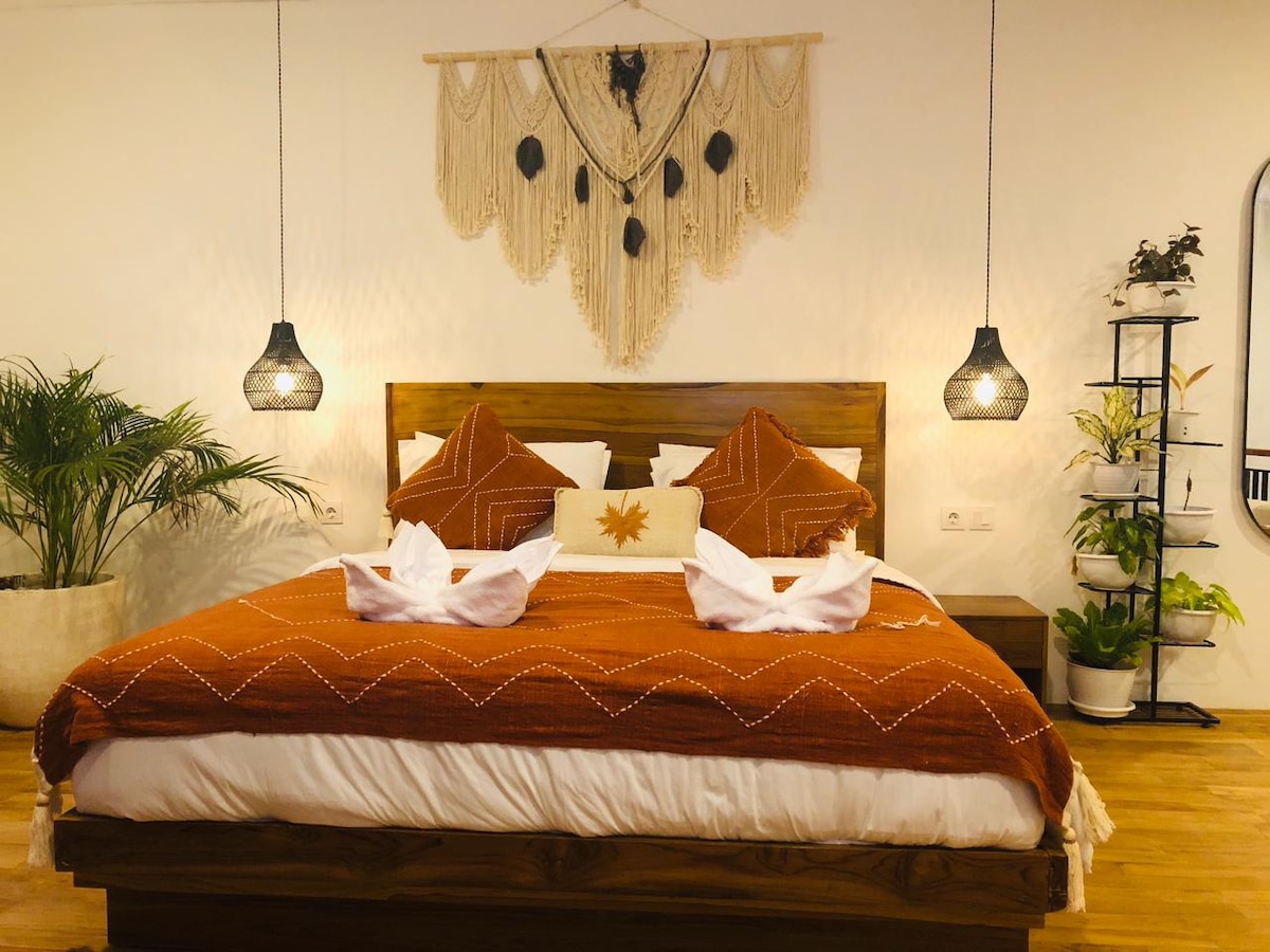 The bedroom features a spacious bed adorned with an earthy-toned blanket and decorative pillows. Two pendant lights illuminate the area, highlighting a tasteful wall hanging above the bed. A small plant stand with greenery is positioned to one side, adding a natural element.