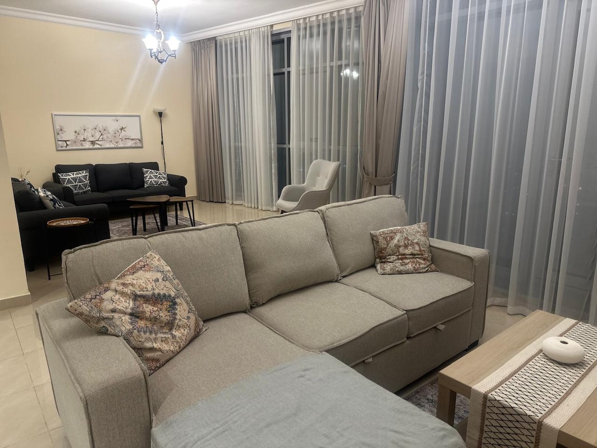 A spacious living area is presented with a light grey sectional sofa, featuring decorative pillows. A wooden coffee table sits nearby, and two armchairs are positioned to one side. Large windows are covered with sheer curtains, allowing natural light to fill the room.