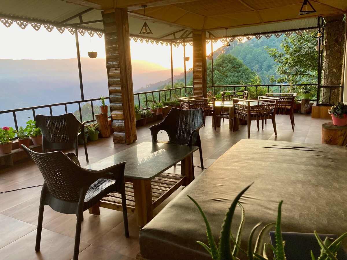 A spacious outdoor terrace features comfortable seating arrangements with wicker chairs and a large table. Potted plants add a touch of greenery. The backdrop showcases a breathtaking view of hills and a sunset, enhancing the tranquil atmosphere.