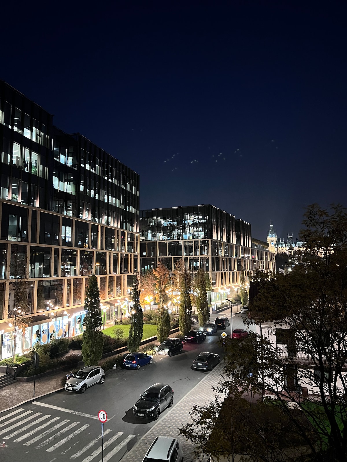 An urban view at night captures modern buildings lined with illuminated windows. Trees provide greenery along the street, where cars are parked and moving. The distant skyline features a prominent tower, contributing to the vibrant city atmosphere.
