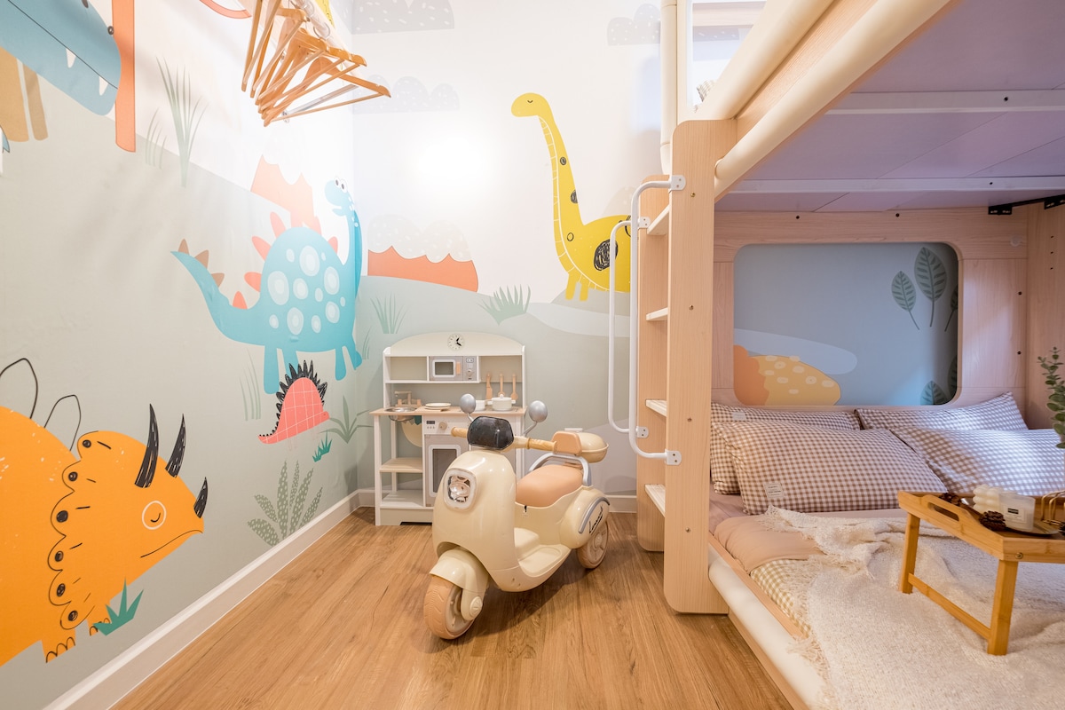 A children's bedroom features a vibrant dinosaur-themed mural on the wall. A cozy sleeping area is equipped with a spacious bed and surrounded by playful decor. A little motorbike toy and a miniature play kitchen are positioned on the wooden floor, adding to the playful atmosphere.