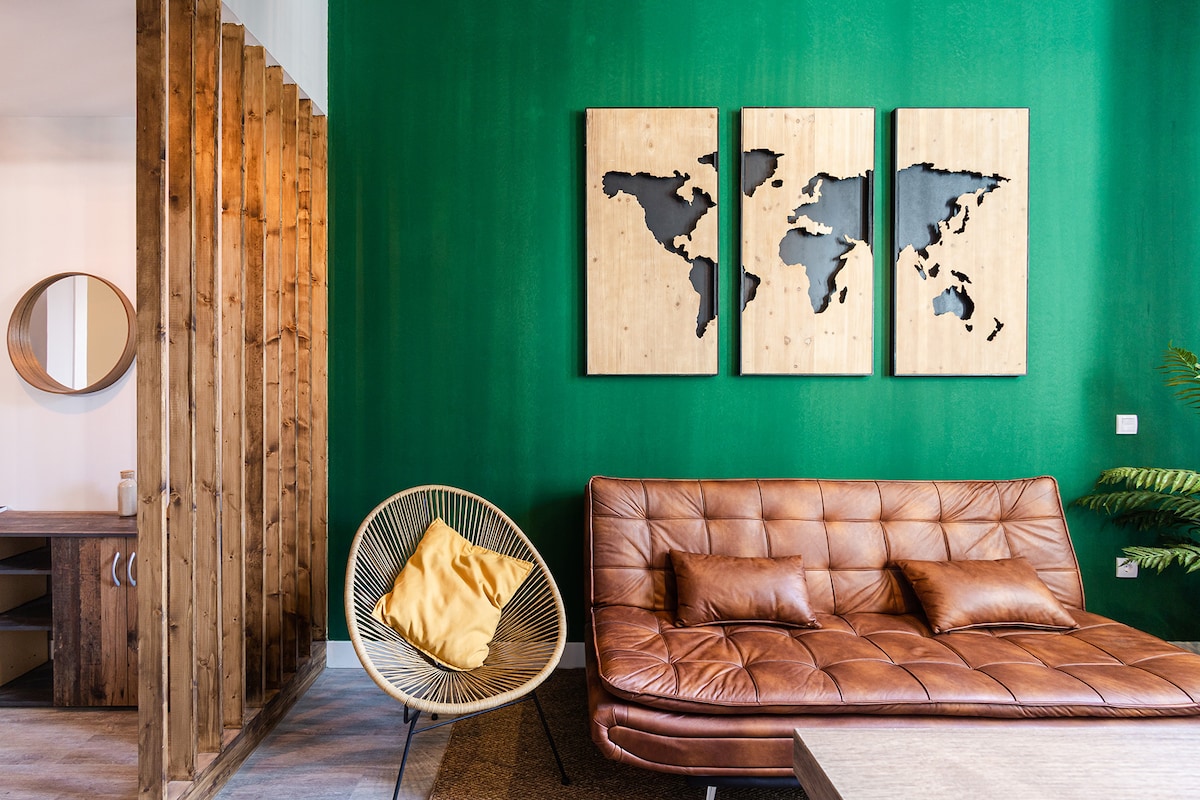 A cozy living area features a green accent wall adorned with three wooden world map panels. A brown leather sofa with cushions is positioned opposite a round mirror and a unique chair with a yellow accent pillow, highlighting a modern yet inviting design.