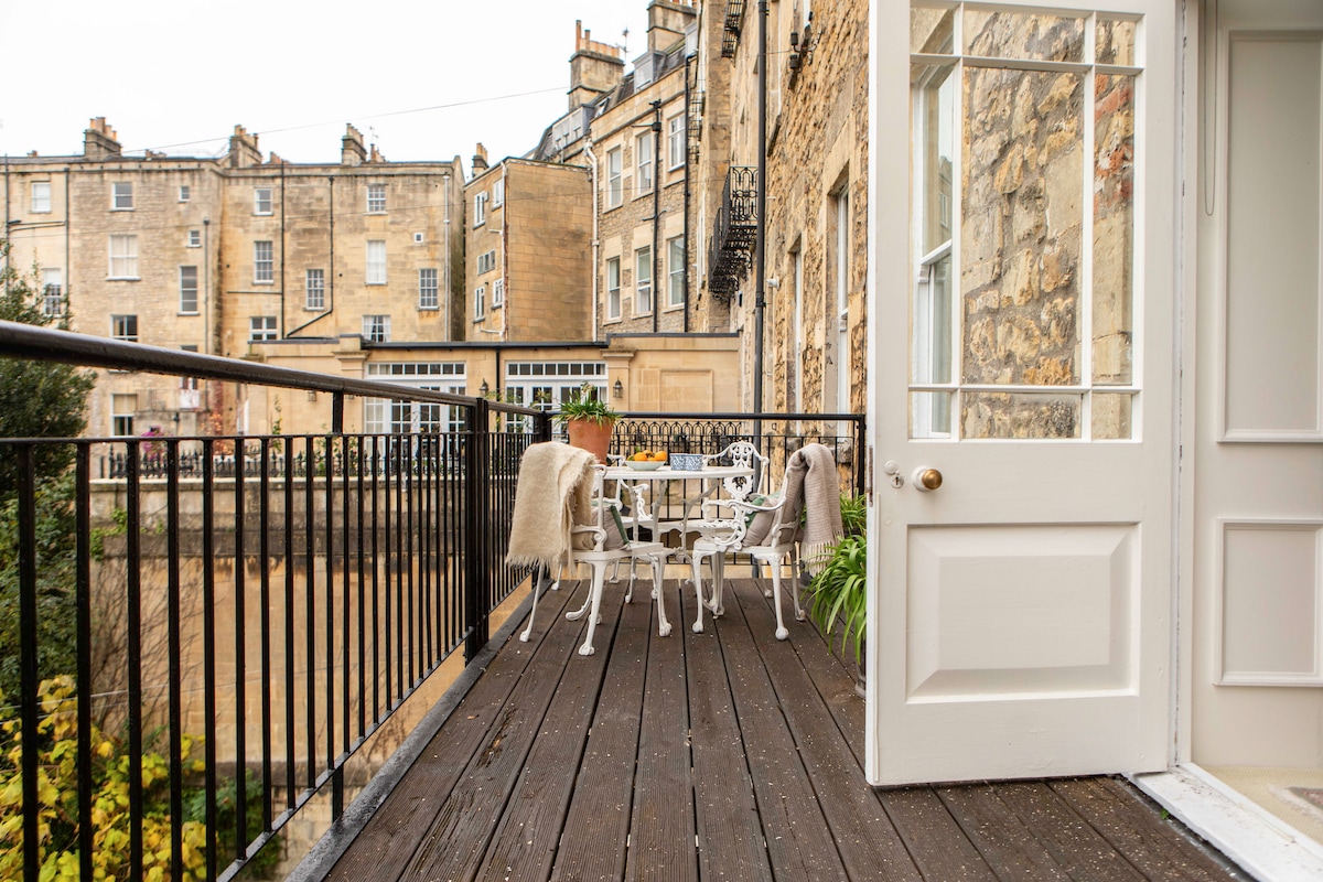 A spacious balcony features wooden flooring with white outdoor furniture, including a table and chairs. Soft blankets drape over the chairs, and potted plants add greenery. The view reveals the historic stone buildings of Bath, creating a serene outdoor retreat ideal for relaxation.