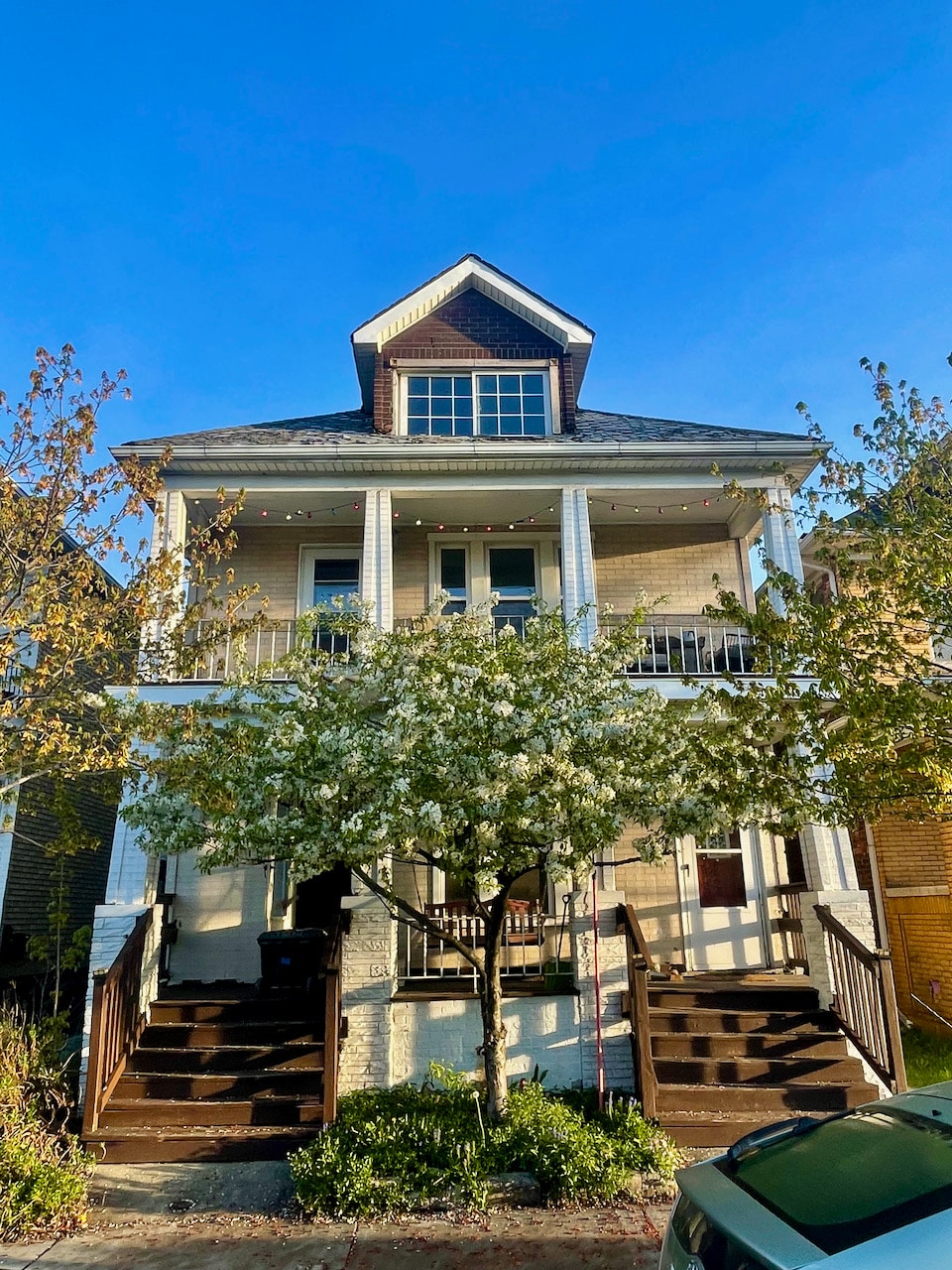 A charming two-story house is presented, featuring a gabled roof and welcoming front porch. Two sets of stairs lead to the entrance, framed by flowering trees. String lights are suspended above the porch, adding to the home’s inviting ambiance. A clear blue sky completes the scene.